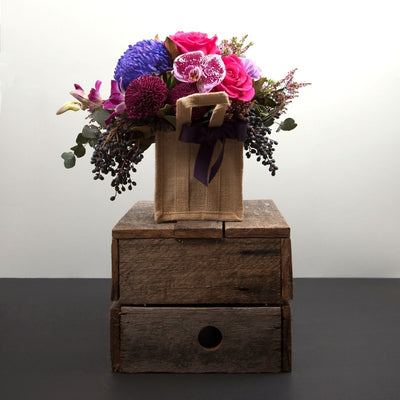 Berry Crush Floral Arrangement in Hessian Bag. Mixture of pink, red, lilac, and burgundy flowers with luscious foliage and leaves.