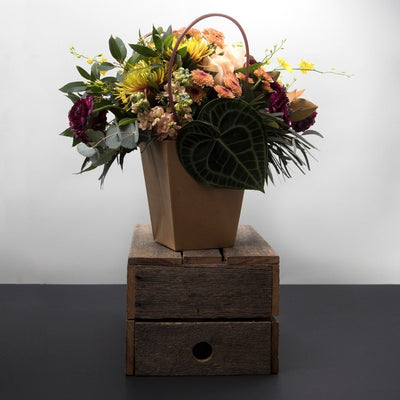 Sunrise Florals in Bag Arrangement. Mixture of beautiful beige, tangerine and yellow flowers with luscious foliage and leaves. Image shown is styled with Dancing Lady Orchids, Turtle Leaves, Roses, Gum Leaves, and more. Available in 3 sizes small, medium, and large. Presented in our exquisite bag arrangement.